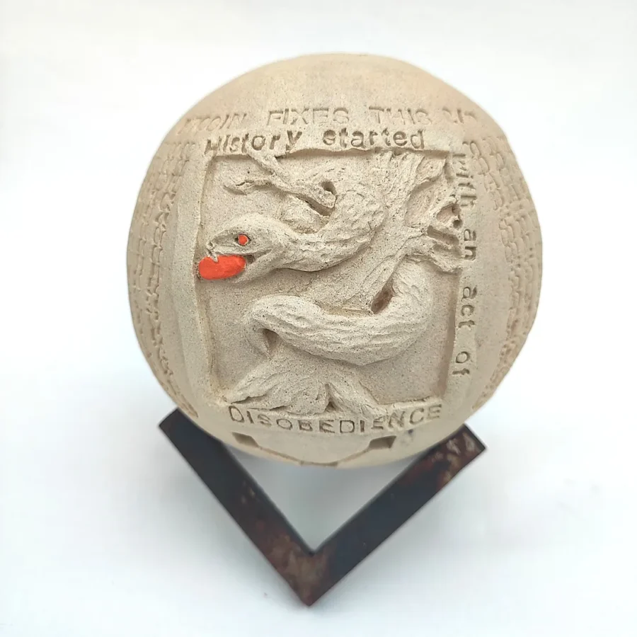 Yonat Vaks Bulla clay artwork of a snake holding a red apple in a tree, inscribed with 'History Started with an act of Disobedience' and 'Bitcoin Fixes This', on a metal stand.